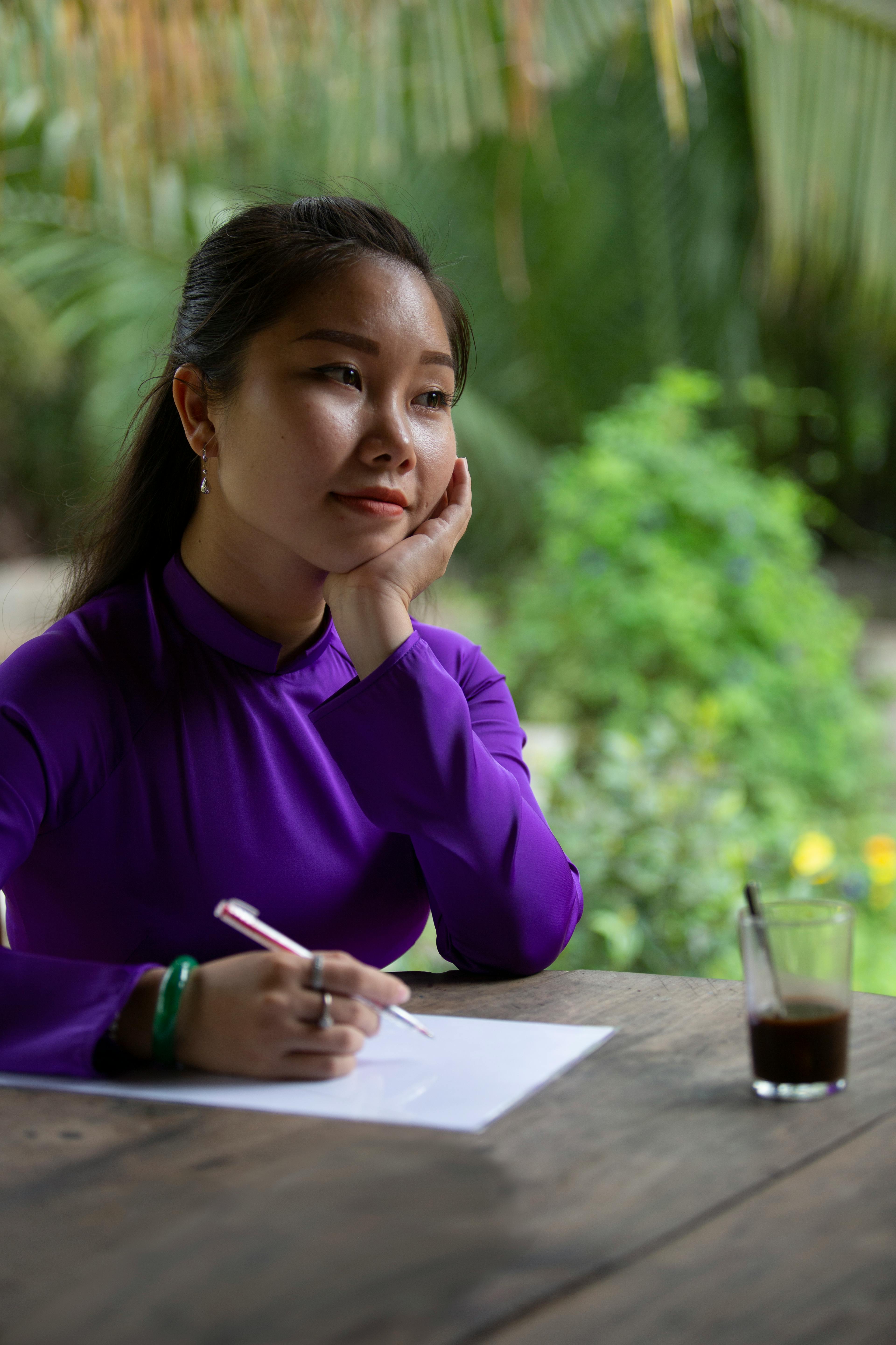 Asian woman in purple pondering with pen and paper at outdoor table.