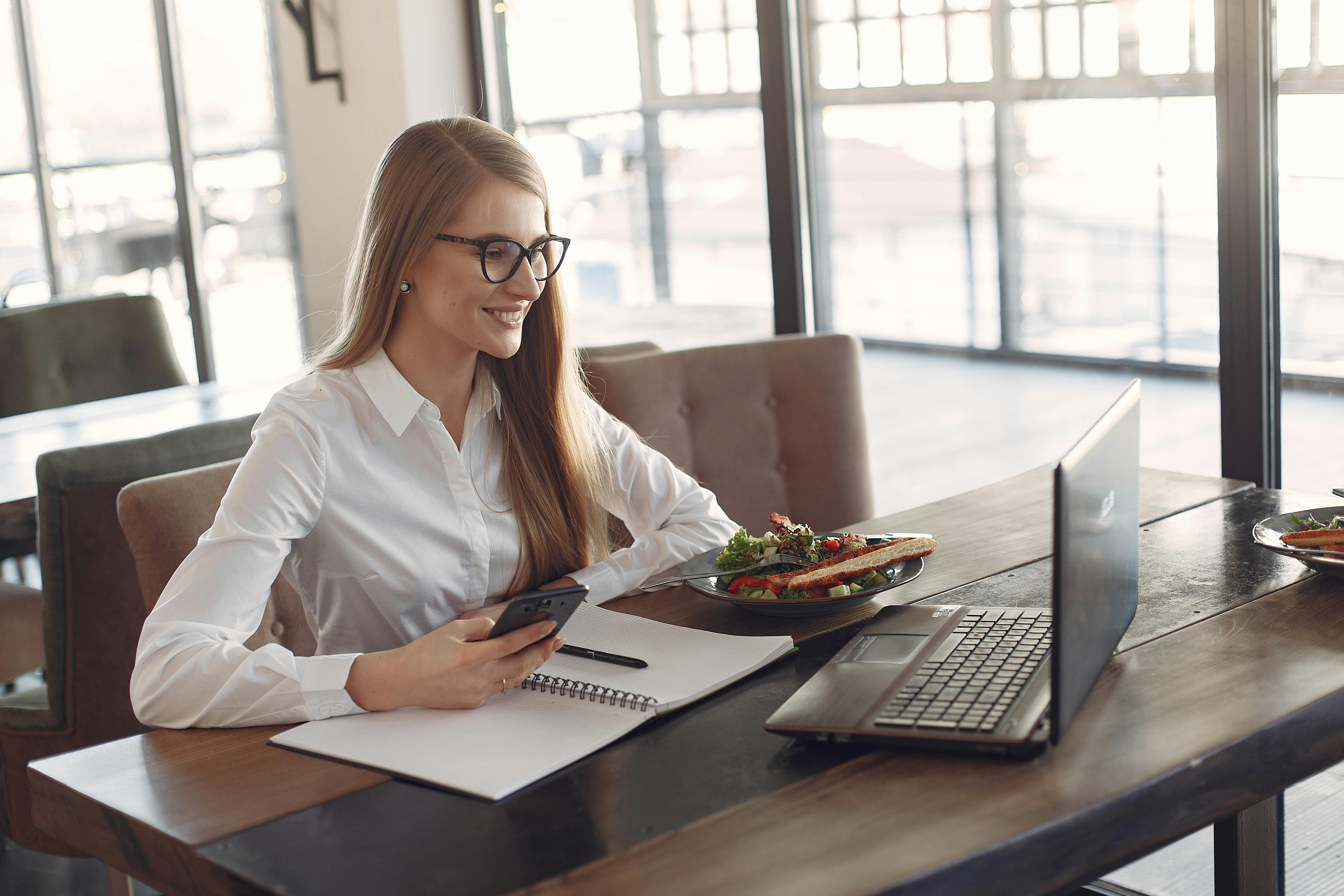 Smiling businesswoman in white shirt using smartphone and laptop at a café while enjoying lunch.